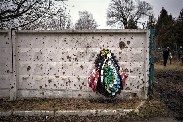 A wreath is photographed leaning against a shrapnel-damaged wall in the cemetery where the funeral for Kasich, 42, is being held, in Bucha, near Kyiv, Ukraine, Friday, February 17, 2023. Kasich Kostiantyn, 42, as senior lieutenant of 93rd Ukrainian brigade was killed on Tuesday, Feb. 14 in the fightings in Bakhmut area. (Photo by Emilio Morenatti/AP Photo)