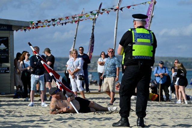 A police officer stands on duty as a protester from the “Enough is Enough” demonstration called by far-right activists lies on the sand with his Union Jack flag in Weymouth, on the southwest coast of England where the Bibby Stockholm migrant accommodation barge is moored, on August 4, 2024. Far-right protesters clashed with British police during tense rallies as unrest linked to disinformation about a mass stabbing that killed three young girls spread across the UK. (Photo by Justin Tallis/AFP Photo)