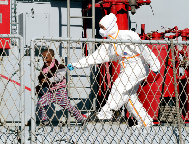 A worker in a sanitary outfit plays with a child on the deck of the Italian Navy frigate Bersagliere as migrants arrive at the Reggio Calabria harbor, Italy, Monday, May 4, 2015. Italy's Coast Guard and Navy as well as tugs and other commercial vessels joined forces to rescue migrants in at least 16 boats Sunday, saving hundreds of them and recovering 10 bodies off Libya's coast, as smugglers took advantage of calm seas to send packed vessels across the Mediterranean. (Photo by Adriana Sapone/AP Photo)