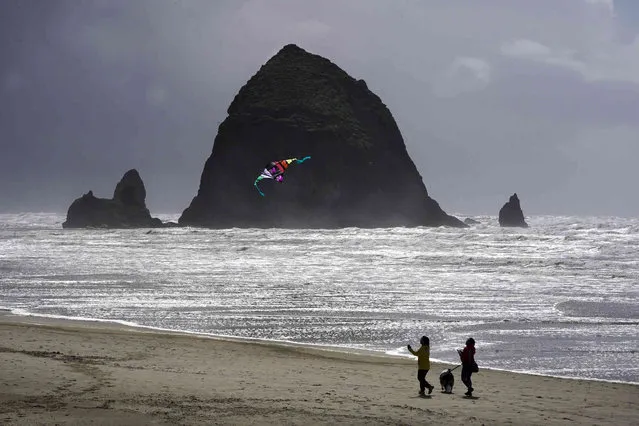 Beachgoers walk a dog and fly a kite as they near Haystack Rock, Monday, April 4, 2022, in Cannon Beach, Oregon. Formed by lava flows from the Blue Mountains and Columbia Basin, it is a popular tourist attraction towering 235 feet and is home to the state's largest Tufted Puffin colony. (Photo by Charles Rex Arbogast/AP Photo)