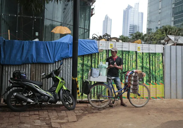 In this Friday, October 14, 2016 photo, a coffee vendor waits for customers near a patch of metal sheets adorned with stickers depicting a forest used to cover a construction site in Jakarta, Indonesia. The capital's fondness for fake greenery creates a n irony as the country is known for cutting down its precious tropical forests at a record rate. (Photo by Dita Alangkara/AP Photo)