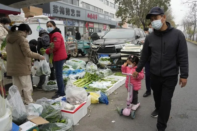 A child wearing a mask passes by residents buying fresh vegetables from street vendors as restaurants are closed in some districts in Beijing, Thursday, November 24, 2022. (Photo by Ng Han Guan/AP Photo)