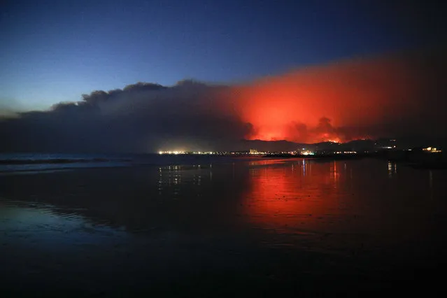 A wildfire continues to burn as its red glow is reflected on the beach Tuesday, December 5, 2017, in Ventura, Calif. Raked by ferocious Santa Ana winds, explosive wildfires northwest of Los Angeles and in the city's foothills burned a psychiatric hospital and scores of homes Tuesday and forced the evacuation of tens of thousands of people. (Photo by Jae C. Hong/AP Photo)