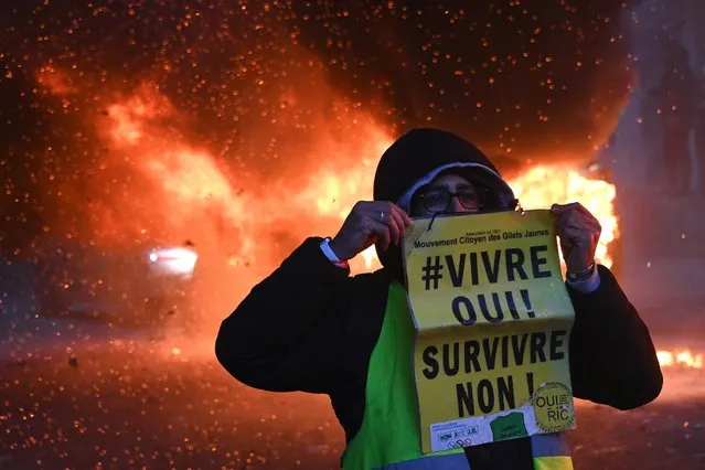 A “Yellow Vest” (Gilet Jaune) anti-government protestor poses with a sign reading “Living, Yes! Surviving, No!” near burning cars during a demonstration for “social rights” and against the “global security” draft law, which Article 24 would criminalise the publication of images of on-duty police officers with the intent of harming their 'physical or psychological integrity', in Paris, on December 5, 2020. (Photo by Anne-Christine Poujoulat/AFP Photo)