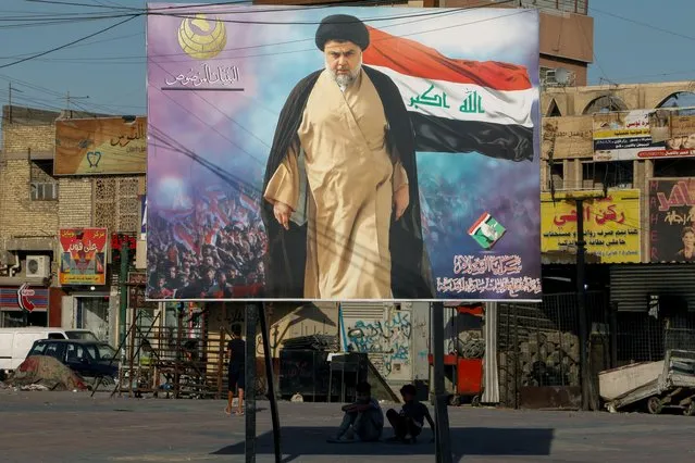 Iraqi boys sit under a poster of Iraqi Shi'ite cleric Moqtada al-Sadr in the Sadr City district of Baghdad, Iraq on June 21, 2021. The text at the top left of the poster reads “The Solid Structure”. The text at the bottom right of the poster reads “Saraya al-Salam, operation command of holy Samarra”. (Photo by Ahmed Saad/Reuters)