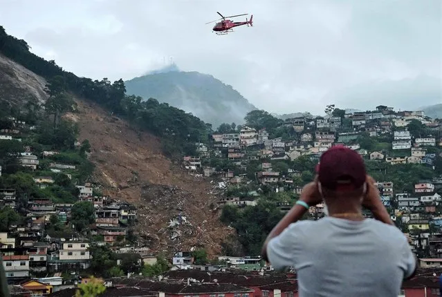 A man takes a picture to an emergency service helicopter flying over a large landslide caused by severe flash floods in Petropolis, Brazil, on February 18, 2022. The Brazilian city of Petropolis is under heavy rains this Friday, three days after a historic storm that left at least 122 dead and covered entire neighborhoods with mud. (Photo by Carl De Souza/AFP Photo)