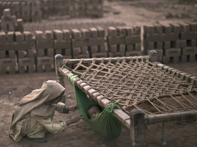 Pakistani girl, Kiran Riasat, 8, who works with her mother and father, seen in the background, in a brick factory, checks on her brother, Rizwan, 1.5, laying in a hammock attached on a bed, at the site of work, in the outskirts of Islamabad, Pakistan, Tuesday, February 18, 2014. According to Kiran's parents she started to work at the factory 3 years ago, and they couldn't afford financially sending her to a school. (Photo by Muhammed Muheisen/AP Photo)