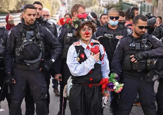 A protester dressed as a clown holds her hand up to the top of her chest where a symbolic heart is placed, as she stands before before Israeli policemen during a demonstration in the flashpoint neighbourhood of Sheikh Jarrah in Israeli-annexed east Jerusalem on February 18, 2022. Hundreds of Palestinians face eviction by Israel from homes in Sheikh Jarrah and other east Jerusalem neighbourhoods. Circumstances surrounding the eviction threats vary. (Photo by Ronaldo Schemidt/AFP Photo)