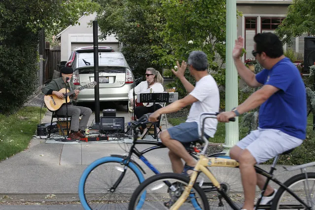 A pair of bicyclists, foreground, wave to musicians George Sheldon, left, and Sandra Carter as they perform a neighborhood concert in Sacramento, Calif., Friday, April 24, 2020. With people following the mandatory stay-at- home directive, the duet was invited by friends to perform a Friday night show. The pair that performs locally by the name “Be Here Now”, has had several appearances canceled due to the coronavirus pandemic. (Photo by Rich Pedroncelli/AP Photo)