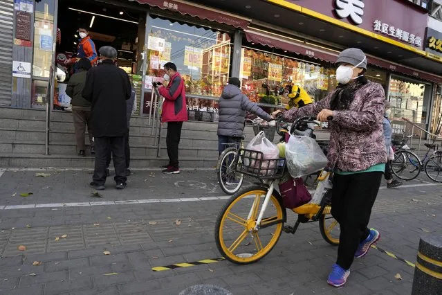 A resident wearing a mask pushes a bicycle with her groceries past a store controlling the flow of shoppers in Beijing, Friday, November 25, 2022. Residents of China's capital were emptying supermarket shelves and overwhelming delivery apps Friday as the city government ordered accelerated construction of COVID-19 quarantine centers and field hospitals. (Photo by Ng Han Guan/AP Photo)