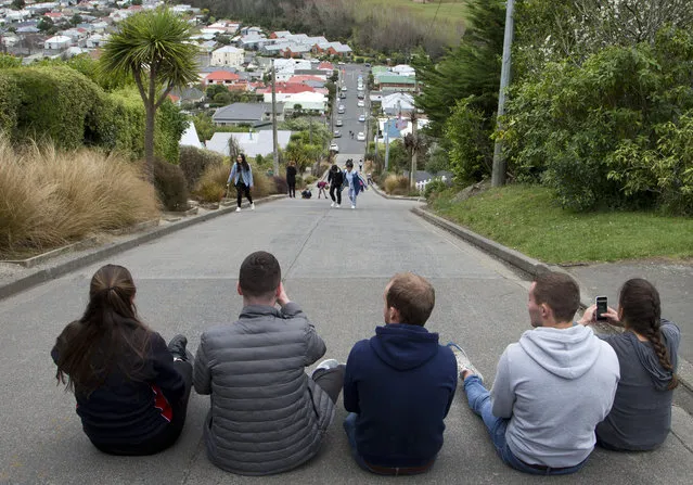 Tourists sit in the middle of the world's steepest street, Baldwin street in Dunedin, New Zealand, Saturday, August 26, 2017. Baldwin Street is recognized by the Guinness Book of Records as the steepest residential street in the world with a the slope of about 1:2.86 (19° or 35%) – that is, for every 2.86 meters travelled horizontally, the elevation rises by 1 meter. (Photo by Mark Baker/AP Photo)