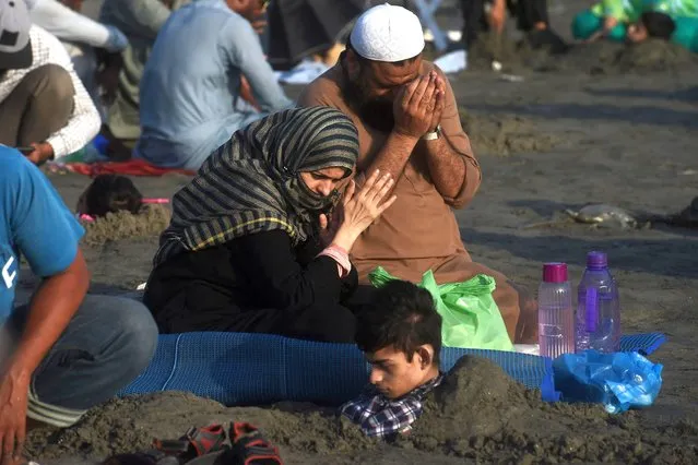 Parents pray next to their son who is covered in sand in a beach with the believe that exposure during a solar eclipse will heal their son illness, in Karachi on October 25, 2022. (Photo by Asif Hassan/AFP Photo)