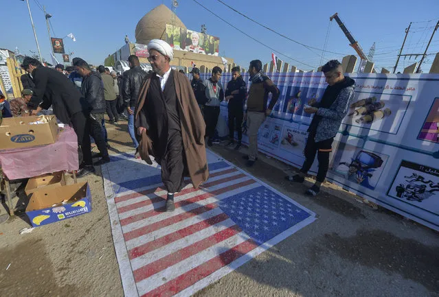 An Iraqi cleric walks on a US flag painted on the ground as people gather to pay respects by the grave of slain commander Abu Mahdi al-Muhandis at the Wadi al-Salam (“Valley of Peace”) cemetery in the holy city of Najaf, on January 4, 2021, marking the first anniversary of his killing alongside Iranian Revolutionary Guards commander Qasem Soleimani in a US drone strike. (Photo by Ali Najafi/AFP Photo)