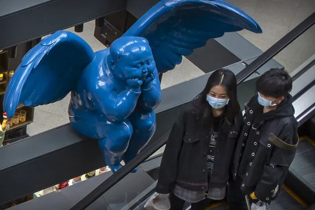 People wearing face masks ride an escalator near a winged statue at an upscale shopping mall in Beijing, Tuesday, March 31, 2020. China on Tuesday reported just one new death from the coronavirus and a few dozen new cases, all brought from overseas. (Photo by Mark Schiefelbein/AP Photo)