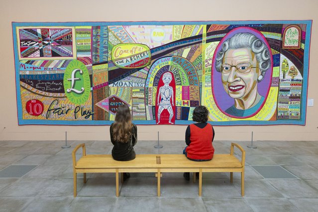 “Comfort Blanket” by Grayson Perry, a colorful tapestry featuring a portrait of Queen Elizabeth II and various British cultural references, with two people sitting on a bench admiring it on October 23, 2025. (Photo by James Glossop/The Times & Sunday Times)