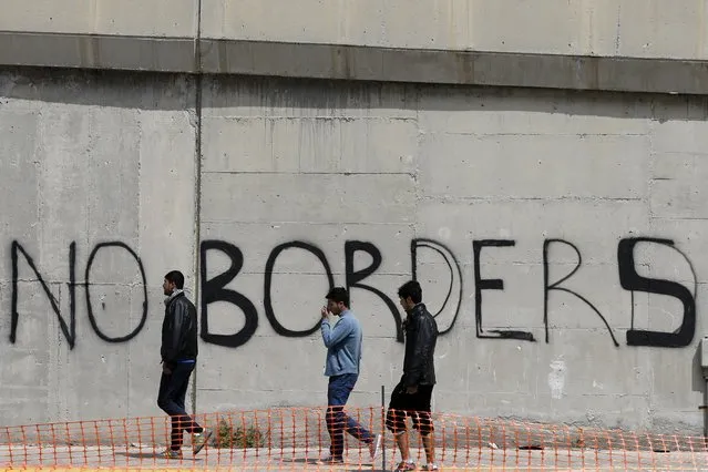 Migrants walk past graffiti at the port city of  Piraeus, Greece on Monday, April 25, 2016. Around 3,500 migrants remain in Piraeus using tents, a warehouse and a terminal passenger building for shelter, as over 50,000 stranded refugees and migrants remain in the country. (Photo by Thanassis Stavrakis/AP Photo)