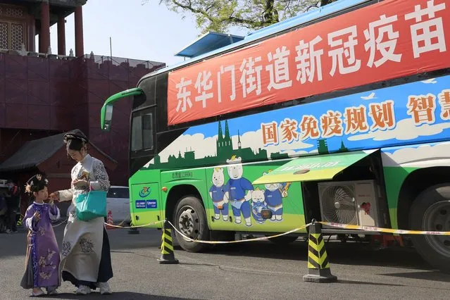 A woman and child wearing period costumes pass by a bus providing COVID-19 vaccination outside the Forbidden City in Beijing on April 14, 2021. If China is to meet its tentative goal of vaccinating 80% of its population against the coronavirus by the end of the year, tens of millions of children may have to start rolling up their sleeves. (Photo by Ng Han Guan/AP Photo)