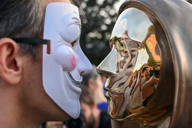 A participant wearing a Guy Fawkes mask poses in front of the statue of Satoshi Nakamoto, the mysterious inventor of the virtual currency bitcoin, after its unveiling at the Graphisoft Park in Budapest, on September 16, 2021. Hungarian bitcoin enthusiasts unveiled a statue on September 16 in Budapest that they say is the first in the world to honour Satoshi Nakamoto, the mysterious inventor of the virtual currency. The bronze life-size sculpture depicts a hooded figure with stylised facial features, alluding to Nakamoto, a pseudonym credited as bitcoin's founder, but whose identity remains unknown. (Photo by Attila Kisbenedek/AFP Photo)