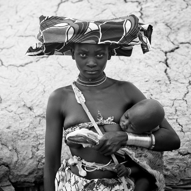 “Mucubal Woman Holding Her Baby, Virie Area, Angola. Mucubal especially women, are famous for the way they dress. The latter wear an original and unique headdress called the Ompota. It is made of a wicker framework, traditionally filled with a bunch of tied cow tails, decorated with buttons, shells, zippers and beads. But tradition is disappearing as some women use barbie dolls boxes to cram their ompota headdress. Women whether they are married or not can wear jewels. Ornaments like iron anklets, called Othivela, and armlets, called Othingo, are worn by girls as well as adult women. Mucubal women are also famous for the string they have around their breast, called oyonduthi, which is used as a bra. Women use to smoke tobacco (that they keep in a snuffbox called boceta) in pipes called opessi”. (Eric Lafforgue)