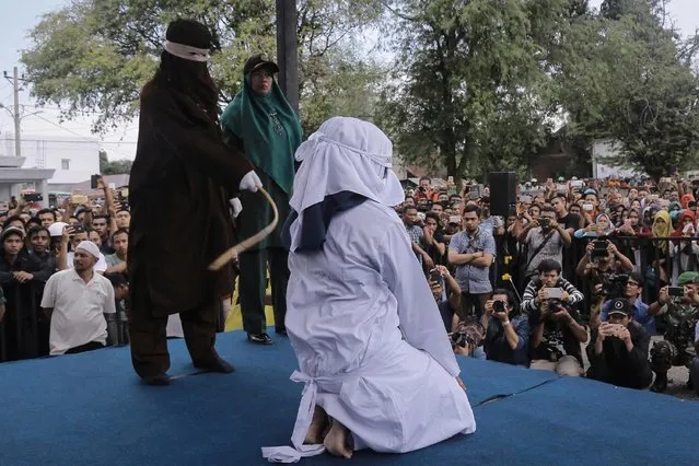 A Shariah law official whips a woman who is convicted of prostitution during a public caning outside a mosque in Banda Aceh, Indonesia, Friday, April 20, 2018. Indonesia's deeply conservative Aceh province on Friday caned several unmarried couples for showing affection in public and two women for prostitution before an enthusiastic audience of hundreds. The canings were possibly the last to be carried out before large crowds in Aceh after the province's governor announced earlier this month that the punishments would be moved indoors. (Photo by Heri Juanda/AP Photo)