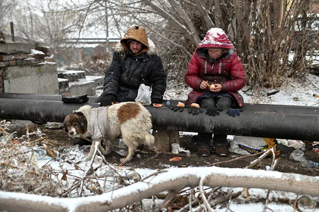 Sasha, 49, nicknamed “Poltorashka” (1,5-litre beverage bottle) and Lyusya Stepanova, 44, both of whom are homeless, sit on a warm pipe with their dog Bim, as they share a meal in Omsk, Russia, December 3, 2019. (Photo by Alexey Malgavko/Reuters)