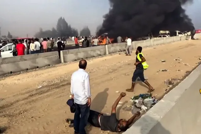 A grab from a UGC video posted on Facebook on October 28, 2023, shows a man lying on the ground as heavy black smoke billows from charred vehicles following a collision on the Cairo-Alexandria desert road near Wadi al-Natrun that reportedly left at least 35 people dead and more than 50 people dead. Traffic accidents are common in Egypt where roads are often in bad repair and the highway code is frequently disregarded. (Photo by -UGC/AFP Photo)