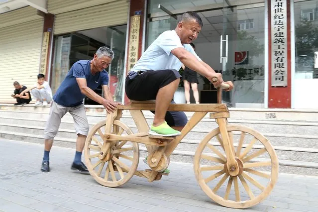He Yong (L) helps a man ride his self-made wooden bicycle in the street on July 12, 2021 in Pingliang, Gansu Province of China. He Yong, 58, made the bicycle without nails, and all wooden components are connected by Sunmao, one of the oldest and most durable structures which were used in Chinese traditional architecture. (Photo by Zheng Bing/VCG via Getty Images)