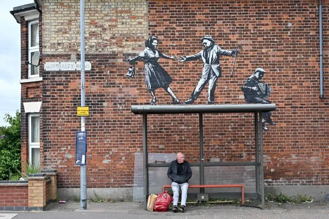 A man waits at a bus stop below a graffiti artwork of a couple dancing to an accordion player, which bears the hallmarks of street artist Banksy, on a wall in Great Yarmouth on the East coast of England on August 8, 2021. The work has not yet been claimed by Banksy. (Photo by Justin Tallis/AFP Photo)