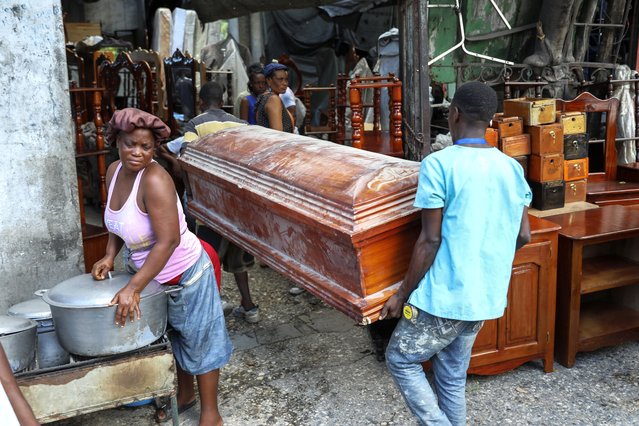 People carry an empty coffin past a street food vendor into the shop that is buying it to repair and sell in Port-au-Prince, Haiti, Friday, September 13, 2024. (Photo by Odelyn Joseph/AP Photo)