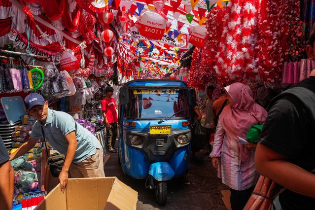 A three-wheeled motorized vehicle drives past people in an alley where vendors have displayed national flags and decoration material for sale ahead of country's Independence anniversary in Jakarta, Indonesia, Tuesday, August 13, 2024. (Photo by Tatan Syuflana/AP Photo)