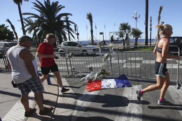 People jod by a French flag and flowers laid near the scene of a truck attack in Nice, southern France, Saturday, July 16, 2016. Nice's seaside boulevard reopens to traffic Saturday following a dramatic truck attack which killed more than 80 people and wounded more than 200 others at a fireworks display. (Photo by Luca Bruno/AP Photo)