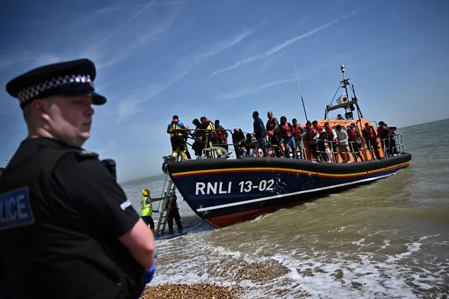 A British police officer stands guard on the beach of Dungeness, on the southeast coast of England, on June 15, 2022, as Royal National Lifeboat Institution's (RNLI) members of staff help migrants to disembark from one of their lifeboat after they were picked up at sea while attempting to cross the English Channel. Furious Conservatives called on Britain's government on June 15, 2022 to abandon a European human rights pact after a judge dramatically blocked its plan to fly asylum-seekers to Rwanda. Under the UK's agreement with Rwanda, all migrants arriving illegally in Britain are liable to be sent to the East African nation thousands of miles away for processing and settlement. The government, after arguing that Brexit would lead to tighter borders, says the plan is needed to deter record numbers of migrants from making the perilous Channel crossing from northern France. (Photo by Ben Stansall/AFP Photo)