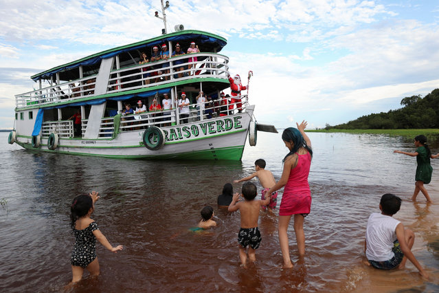 A member of the Amigos do Papai Noel (Friends of Santa Claus group) dressed in a Santa Claus costume greets residents from a boat during a visit to the community located along the Negro River, in Iranduba, Amazonas State, Brazil on December 17, 2022. (Photo by Bruno Kelly/Reuters)