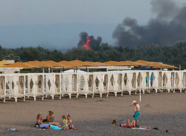 People rest on a beach as smoke and flames rise after explosions at a Russian military airbase, in Novofedorivka, Crimea on August 9, 2022. (Photo by Reuters/Stringer)