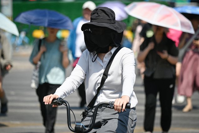 A woman wearing a sun protection mask rides a bicycle across a road on a hot day in Beijing on June 18, 2024. (Photo by Wang Zhao/AFP Photo)