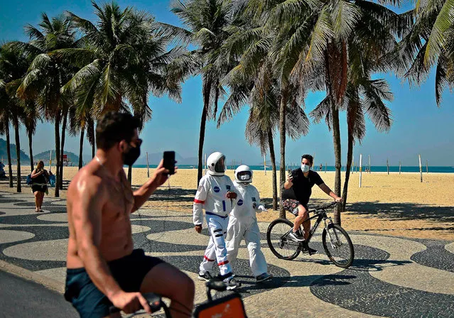 Men riding bikes take pictures of Brazilian accountant Tercio Galdino, 66, and his wife Ailicea who walk along Copacabana beach wearing protective suits, in Rio de Janeiro, Brazil on July 11, 2020. Galdino The has a chronic lung disease, and made the astronaut costumes at home using medical suits used by health professionals. He says that although they allow him protection against coronavirus they are mostly for fun as he has huge interest in Astronomy. (Photo by Carl de Souza/AFP Photo)