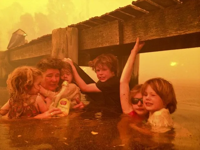 In this January 4, 2013, photo provided by the Holmes family, Tammy Holmes and her grandchildren take refuge under a jetty as a wildfire rages near them  in Dunalley,Australia, on January 9, 2013. The family credits God with their survival from the fire that destroyed around 90 homes there. (Photo by Tim Holmes)