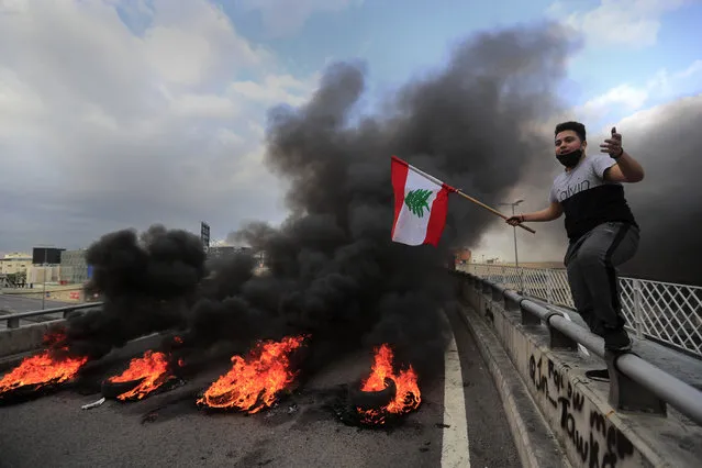 A protester waves a Lebanese flag near burning tires set to block a main highway, during a protest in the town of Jal el-Dib, north of Beirut, Lebanon, Monday, March 8, 2021. The dayslong protests intensified Monday amid a crash in the local currency, increase of consumer goods prices and political bickering between rival groups that has delayed the formation of a new government. (Photo by Hussein Malla/AP Photo)