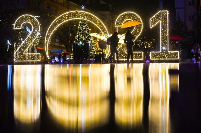 Women walk under an umbrella in front of a 2021 sign displayed in downtown Pristina on December 30, 2020, as Kosovars prepare to celebrate the New Year 2021 at their homes, amid the ongoing Covid-19 (novel coronavirus) pandemic. (Photo by Armend Nimani/AFP Photo)