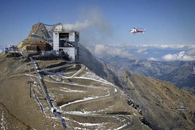 A helicopter carries water to stop an ongoing fire at the restaurant Botta at the Glacier 3000 resort in Les Diablerets, Ormont-Dessus, Switzerland, 19 September 2022. A fire broke out early on 19 September, at a mountaintop restaurant designed by Swiss architect Mario Botta in the Swiss Alps at an altitude of 3,000 metres and helicopters were helping to battle the blaze, police said. (Photo by Laurent Gillieron/EPA/EFE)