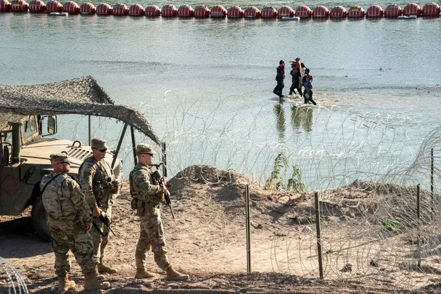 Asylum-seeking migrants walk in the Rio Grande river between a floating fence and the river bank as they look for an opening in a concertina wire fence to land on U.S. soil in Eagle Pass, Texas, U.S. July 24, 2023. (Photo by Go Nakamura/Reuters)