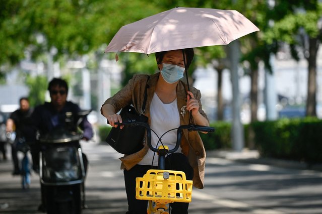 A woman uses an umbrella to shelter from the sun as she rides a bicycle along a street on a hot day in Beijing on June 18, 2024. (Photo by Wang Zhao/AFP Photo)
