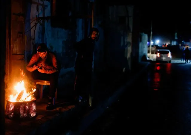 A Palestinian sits by a fire warming himself as a man looks on, on a rainy cold night at Beach refugee camp in Gaza City on January 20, 2022. (Photo by Mohammed Salem/Reuters)
