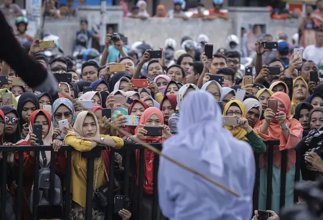 People use their mobile phones to take pictures as a Shariah law official whips a woman who is convicted of prostitution during a public caning outside a mosque in Banda Aceh, Indonesia, Friday, April 20, 2018. Indonesia's deeply conservative Aceh province on Friday caned several unmarried couples for showing affection in public and two women for prostitution before an enthusiastic audience of hundreds. The canings were possibly the last to be carried out before large crowds in Aceh after the province's governor announced earlier this month that the punishments would be moved indoors. (Photo by Heri Juanda/AP Photo)