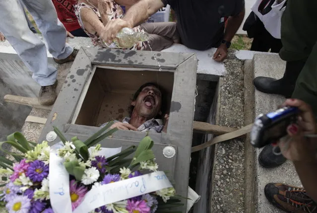 In this February 5, 2014 photo, Divaldo Aguiar, who plays the part of Pachencho, lies inside a mock coffin as villagers splash rum into Aguiar's mouth during the Burial of Pachencho celebration at a cemetery in Santiago de Las Vegas, Cuba. The bash kicked off Wednesday with the slow procession to the local cemetery. Pallbearers carried the coffin of “Pachencho”, who's known the other 364 days of the year as Divaldo Aguiar, to an open grave and used ropes to lower it six feet under. (Photo by Franklin Reyes/AP Photo)