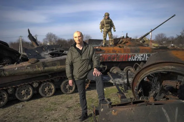 A man poses for the picture wearing a mask of Russian President Vladimir Putin, while a Ukrainian soldier stands on top of a destroyed Russian tank in Bucha, on the outskirts of Kyiv, Ukraine, Thursday, April 7, 2022. (Photo by Rodrigo Abd/AP Photo)