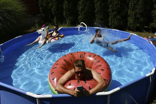 Barnabas types on his phone as he works from the home office as a pool in the garden while his wife, Rebeka, left and sister-in-law, Emma, cool down in Budapest, Hungary, Thursday, August 4, 2022. Hungary has experienced several heat waves since mid June with day-time temperatures rising to 40 degrees Celsius and remaining at tropical levels through the night. In fact, the country recently shattered record for its hottest night ever when temperature of 25.4 C was measured in Budapest on July 26. (Photo by Anna Szilagyi/AP Photo)