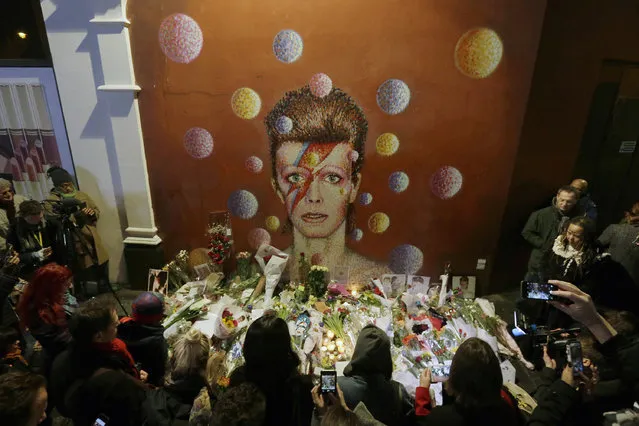 People gather next to tributes placed near a mural of British singer David Bowie by artist Jimmy C, in Brixton, south London, on Jan. 11, 2016. Bowie, the other-worldly musician who broke pop and rock boundaries with his creative musicianship, nonconformity, striking visuals and a genre-spanning persona he christened Ziggy Stardust, died of cancer at age 69. He was born in Brixton. (Photo by Tim Ireland/AP Photo)