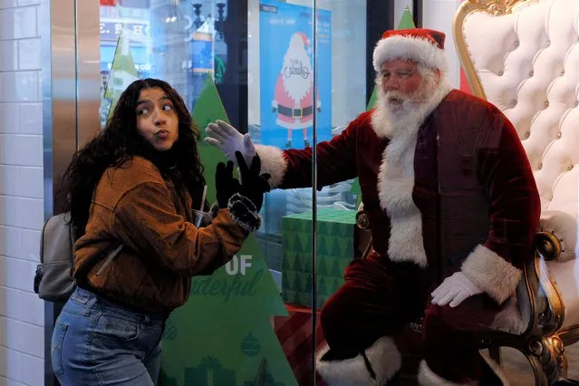 A shopper poses for a photograph with a man in a Santa Claus costume, who is sitting inside a store display window behind glass as a measure to stop the spread of the coronavirus disease (COVID-19) pandemic, during the Christmas holiday season at the Primark store in Boston, Massachusetts, U.S., December 18, 2021. (Photo by Brian Snyder/Reuters)