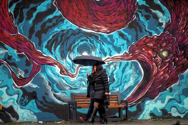 Women shelter under an umbrella as they walk past a wall covered with a mural in Pristina on November 21, 2022. Heavy rain is causing floods in many cities in Kosovo, damaging agricultural land and family businesses Ð while some cities also facing a shortage of drinking water and are suspending school classes. (Photo by Armend Nimani/AFP Photo)